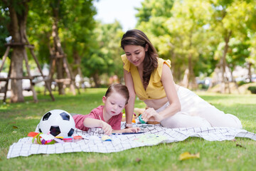 Woman and child sit on picnic blanket in park, coloring with art supplies, surrounded by greenery...