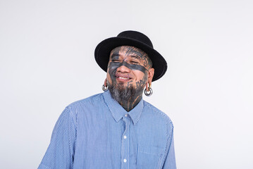 Portrait of a cheerful Asian man fully covered in facial tattoos, showcasing eye tattoos and diverse body piercings, dressed in a stylish hat and striped shirt against a white backdrop.