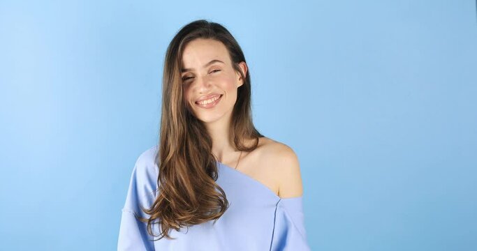 Playful happy flirt brunette woman coquettish blinking eyes, looking at camera with smile and flirting, expressing optimism isolated on blue background. Woman wear blue long sleeves top.