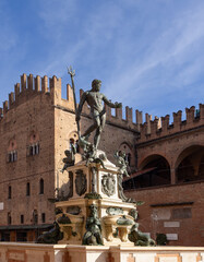 Water jets dance around the bronze Neptune at Bologna fountain, set before the historical Palazzo del Podesta under a blue sky in this vertical image