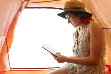 A woman is sitting in a tent reading a book. She is wearing a straw hat and a necklace