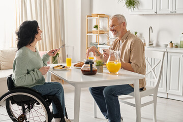 A disabled woman in a wheelchair and her husband sit at a table, enjoying breakfast in their home kitchen.