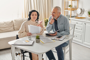A man and a disabled woman in a wheelchair sit at a table in their kitchen at home, planning budget