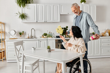 man affectionately gives a bouquet of flowers to his wife in a wheelchair in their kitchen at home.