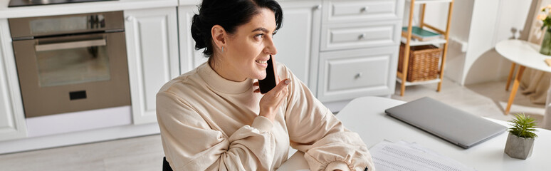 A brunette woman talks at phone and working remotely at a table in her kitchen.