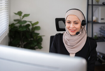 Call center worker, young Muslim woman wearing hijab, talking to customer on call phone on computer...