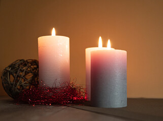 Three candles are lit and placed on a table. The candles are of different sizes and colors, with one being white and the other two being gray. The candles are surrounded by a red and gold decoration
