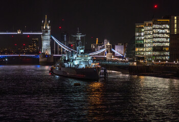 Fototapeta na wymiar Front view of the war museum ship HMS Belfast docked on the River Thames with reflections of lights from restaurants and buildings in the water in front of Tower Bridge illuminated at night.