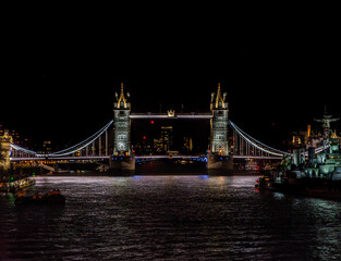 Fototapeta na wymiar London bus driving past Tower Bridge illuminated at night with illuminated skyscrapers and the war museum ship HMS Belfast docked on the River Thames with reflections of restaurant lights in the water