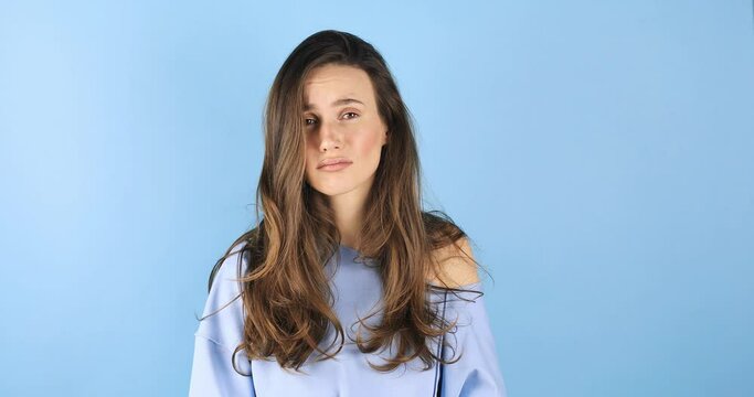 Upset curly brunette hair woman wear blue pullover purses lips offendedly and looks from under forehead on blue background. Sad tired woman looks with poker face at camera purses lip.