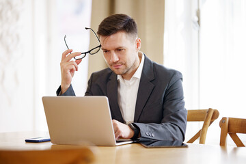 A contemplative businessman pauses, glasses in hand, absorbed in the analysis on his laptop in a...