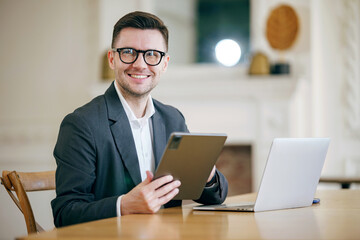An amiable businessman in spectacles expertly coordinates work on his tablet and laptop in a chic,...