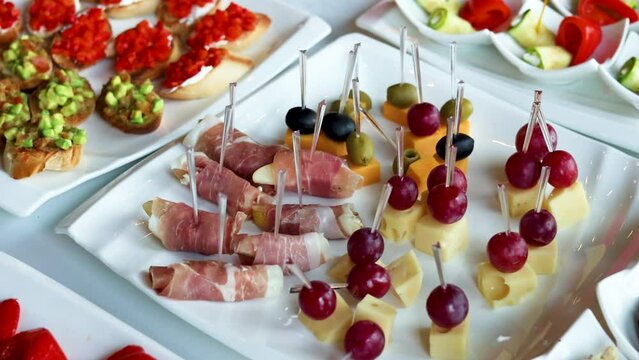 fourchette table with many plates with food.cheese, grape, salami and bread on stick.mini sandwich with salmon, baked pepper and fresh cucumber.many unknown people in background.party, event