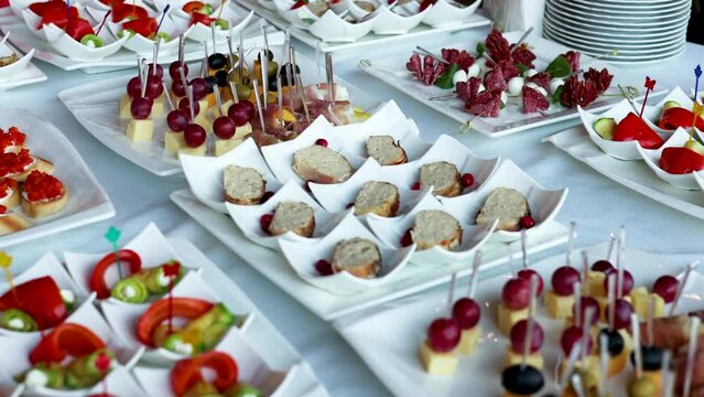 fourchette table with many plates with food.cheese, grape, salami and bread on stick.mini sandwich with salmon, baked pepper and fresh cucumber.many unknown people in background.party, event