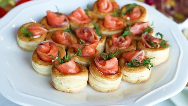 fourchette table with many plates with food.cheese, grape, salami and bread on stick.mini sandwich with salmon, baked pepper and fresh cucumber.many unknown people in background.party, event