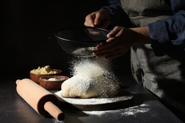 Woman sprinkling flour over dough at black table, closeup