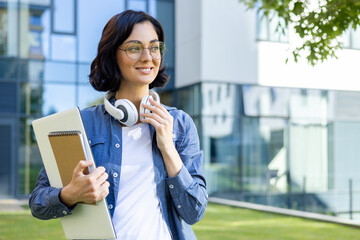 A young smiling female student stands on the street in headphones, holds a laptop and notebooks in...