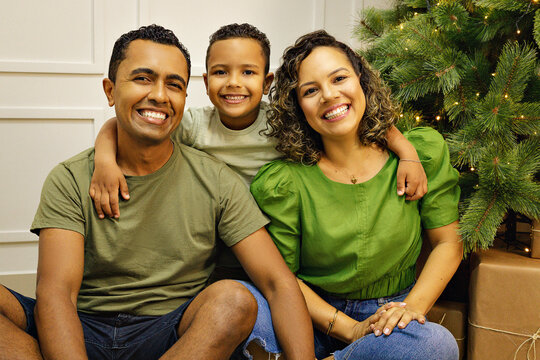 Familia Parda com tra&ccedil;os brasileiros com homem, mulher e um menino crian&ccedil;a, em uma sala com uma decora&ccedil;&atilde;o de natal na cor verde.