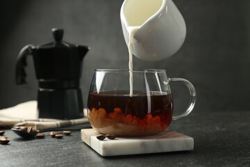 Pouring milk from pitcher into glass cup with coffee at dark textured table, closeup - obrazy, fototapety, plakaty