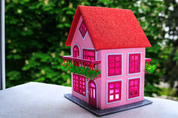 Against a background of green trees, a pink house with a red roof and pink light in the windows