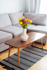 Ceramic Vase with multicolor various flower bouquet on the vintage style wooden coffee table with blurred background of modern cozy light living room with gray couch sofa. Cozy home interior design