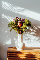 Scandinavian home interior with with exotic protea flowers bouquet in ceramic vase standing on wooden cabinet under sunlight and shadows on white gray wall. Minimalist design of home decor. Vertical