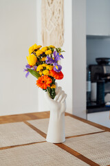 Kitchen counter table with focus on vase made in shape of human hand with multicolor various summer flower bouquet with blurred background of modern cozy white kitchen. Home interior design details.