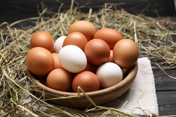 Fresh chicken eggs in bowl and dried hay on black wooden table