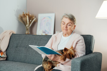 Elderly retired senior woman with wrinkles smiling while embracing her Yorkshire terrier dog pet and relaxing while reading on sofa at home. Best friend. Enjoying retirement lifestyle.