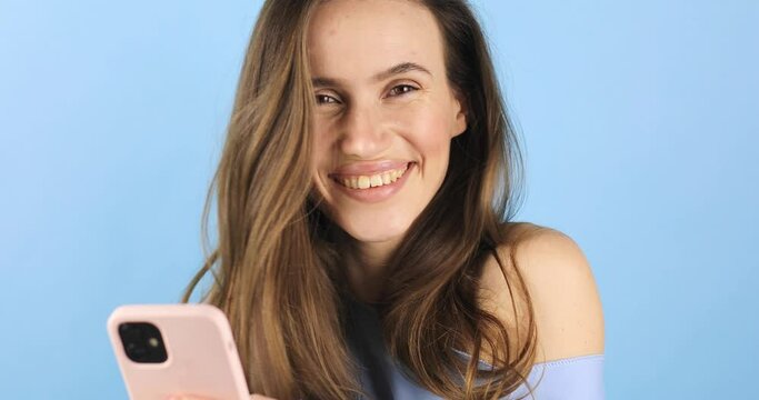 Close up smiling beautiful young woman girl in blue top messaging on smart phone, using mobile cell phone typing browsing chatting send sms on blue background in studio. Girl next look at camera.