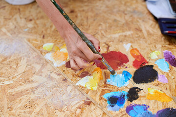 Female painter hand dips paintbrush into palette of colorful paints for live painting of picture for outdoor street exhibition, close up view of female artist hand holding paintbrush