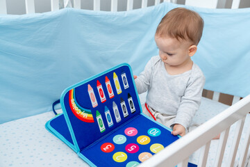 Baby sitting in a crib playing with montessori busy book. Concept of keeping children from screen by activity books and quiet books