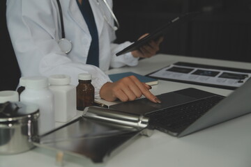 Serious female doctor using laptop and writing notes in medical journal sitting at desk. Young woman professional medic physician wearing white coat and stethoscope working on computer at workplace.