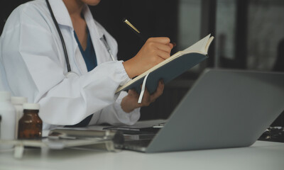 Serious female doctor using laptop and writing notes in medical journal sitting at desk. Young...