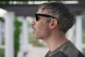 Profile view of a man in his 30s, wearing sunglasses in the park at sunset. The white pergola columns in the background. 