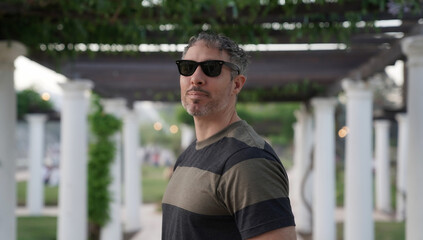 Portrait of a man in his 30s, wearing sunglasses in the park at sunset. The white pergola columns...