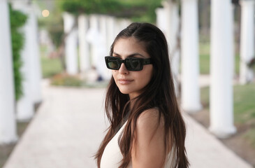 Portrait of a young latin american woman wearing sun glasses, in the park with a beautiful sunset light. The white pergola columns and plants in the background.