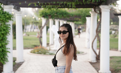 Portrait of a young latin american woman wearing sun glasses, in the park with a beautiful sunset light. The white pergola columns and plants in the background.