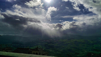 Dramatic sky with sunbeams piercing through clouds over a hilly landscape, casting shadows and...