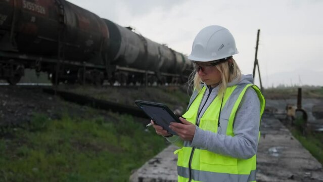 Female railway worker controller wearing white hard hat and safety vest at outdoors near railroad cars, takes notes in digital tablet in wind, background. Copyspace.
