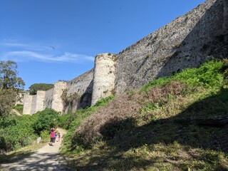 A woman and a child walk on a path in front of a stone wall. The sky is clear and the sun is...