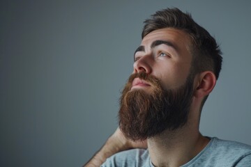 Obraz premium Handsome young bearded man is keeping hand on beard and looking away on a gray background