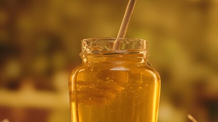 Opened glass jar standing on the table with fresh golden honey, hand dipping honey dipper in the jar with thick syrup, close up shot.