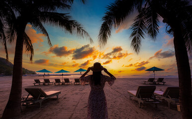 A woman in a hat stands and watches the sunrise on the beach,Young woman in straw hat and a dress standing alone on empty sand beach at sunset sea shore. Lonely girl looking at horizon over calm ocean