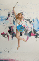 sexy beautiful pretty young artist woman female painter with blond hair in white casual underskirt paints with Brush, paint bucket and Color palette jumps for joy in front of painted studio wall