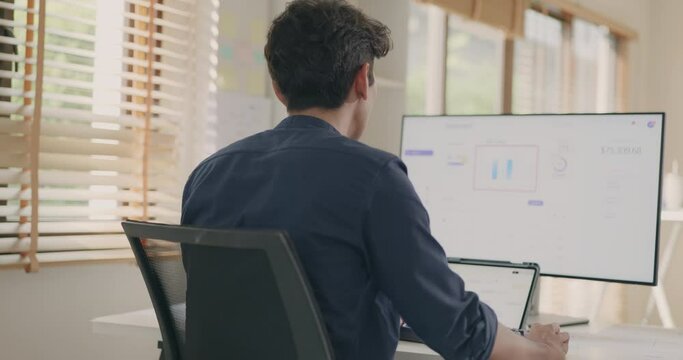 Back view of a young businessman working at his office desk, scrutinizing financial charts on a computer monitor with focus and determination.