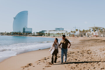 Joyful couple running hand in hand along a sunlit beach in Barcelona, Spain