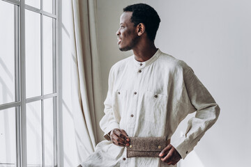 Thoughtful African-American man in casual linen shirt stands by window, holding handmade felt clutch.