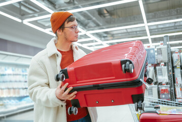 Man Carrying a Red Hardshell Suitcase in a Store, Contemplating Purchase. A young man thoughtfully considers a red suitcase at a store, contemplating its features and suitability for his travel needs