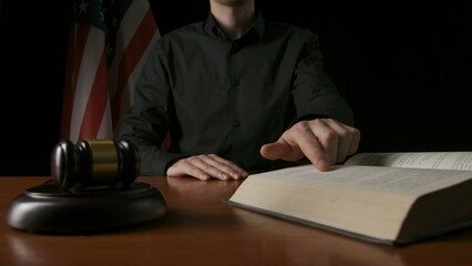 Male sitting at the desk with wooden hammer and law book, USA flag at the background. Man judge in...
